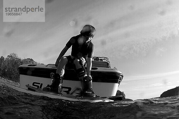 Tiefwinkelperspektive eines kleinen Jungen  der auf dem Heck eines Bootes ein Wakeboard aufsetzt.