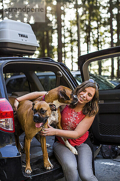 A female owner sits in the back of her vehicle as she allows two dogs to lick her face.