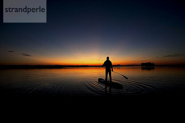 Silhouette eines Mannes beim Stand-up-Paddeln im Abendlicht mit Bäumen  die sich im Wasser spiegeln.