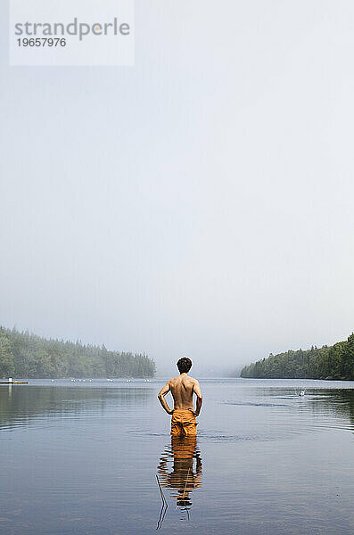 An einem bewölkten Tag steht ein junger Mann im Badeanzug und blickt auf einen kleinen See.