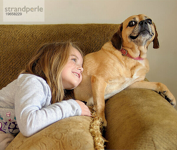 Kleines Mädchen lächelt mit Puggle-Hund auf der Couch