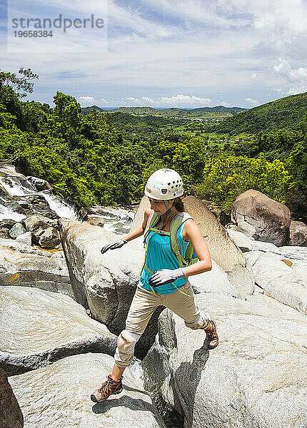 Eine junge Frau hüpft zwischen Felsbrocken herum  während sie an einem sonnigen Tag in Puerto Rico einen Fluss erkundet.