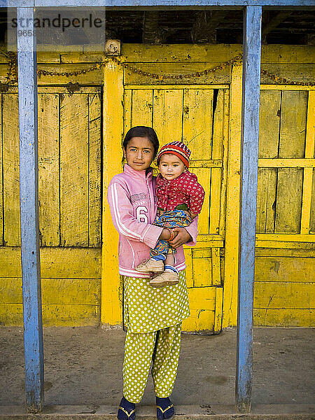 Ein junges nepalesisches Mädchen hält ein kleines Kind vor einer Tür in Nepal.