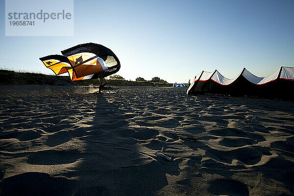 Silhouette eines Mannes  der bei Sonnenuntergang mit einem Kiteboard-Drachen am Strand läuft.