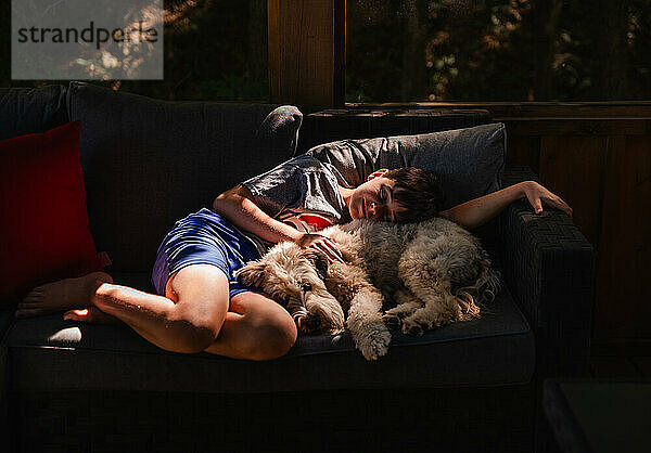 Junge und pelziger Hund liegen zusammen im sonnigen Plätzchen auf dem Sofa.