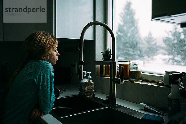 Jugendliches Mädchen beugt sich über Küchenspüle und blickt aus dem Fenster auf Schnee