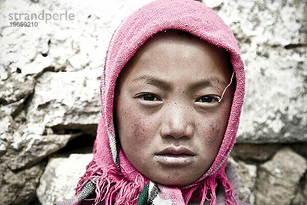 A young school girl with wind burnt cheeks and a pink wrap covering her hair.