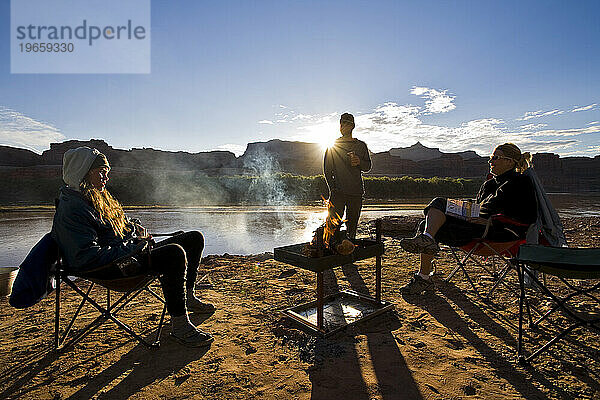Drei Menschen versammelten sich bei Sonnenaufgang um ein Feuer an einem Sandstrand des Colorado River in Utah.