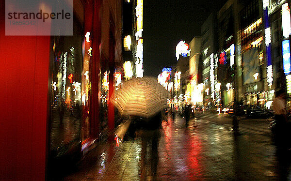 Eine Frau läuft nachts mit einem Regenschirm durch die nassen Straßen Tokios. Shinjuku  Tokio  Japan. (Bewegungsunschärfe) (Unscharf)