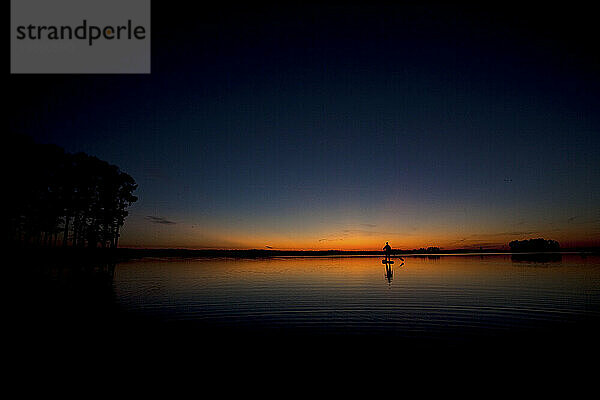 Silhouette eines Mannes beim Stand-Up-Paddleboarden im Sonnenaufgangslicht mit Bäumen  die sich im Wasser spiegeln.