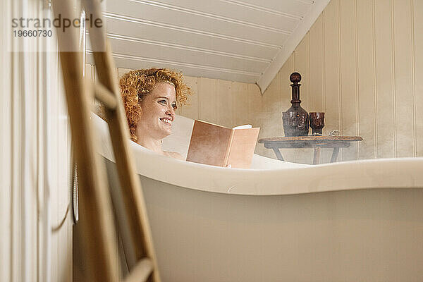 Fröhliche junge Frau liegt in der Badewanne und liest bei Tageslicht ein Buch