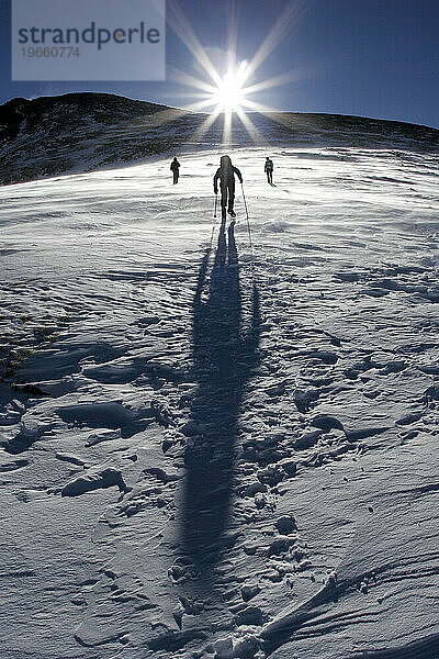 Silhouetten von drei Menschen beim Bergsteigen/Aufsteigen eines Berges in die Sonne.