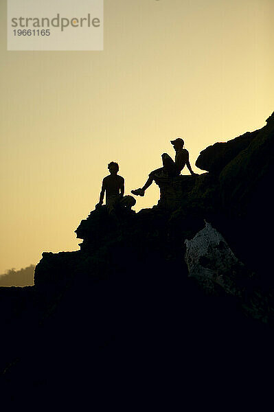 Silhouette zweier Jungen  die auf Felsen in Costa Rica sitzen.