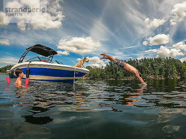 Mann springt an einem Sommertag vom Boot in den See  während Hund und Kind zuschauen.