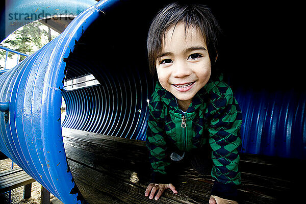 Ein kleiner Junge lächelt  während er durch den Tunnel eines Parks kriecht.