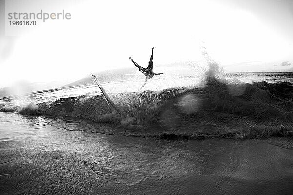 Schwarz-Weiß-Bild eines Skimboarders  der von einer Welle überrollt wird.