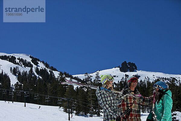 Drei Freunde (ein Mann und zwei Frauen) hängen mit Skiern und einem Snowboard vor einem Skigebiet herum.