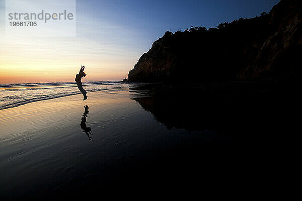 Weibliche Silhouette springt in einen Sonnenuntergang an einem von Klippen umgebenen Strand.