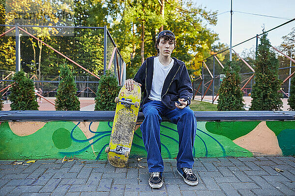 Teenager mit Skateboard sitzt auf einer Bank und blickt in die Kamera