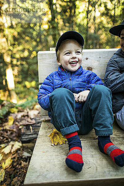 Kleiner Junge sitzt lächelnd auf einer Parkbank und hat gemütliche Kleidung.