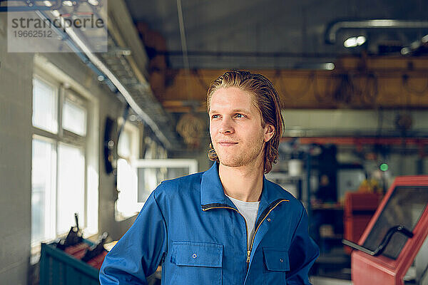 Glücklicher junger Mann in Uniform  der bei Tageslicht in einer verschwommenen Fabrik steht