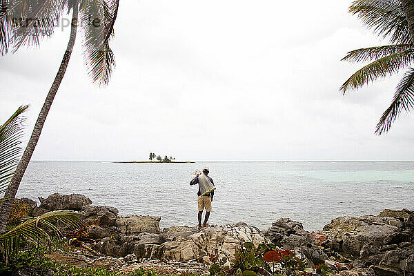Ein Fischer mit einem Netz steht an einem bewölkten Tag am Rand von Felsen mit Blick auf das Meer.