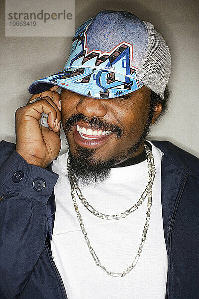 Junger afroamerikanischer Hip-Hop-Mann mit Mütze lächelt  während er ein Handy telefoniert.