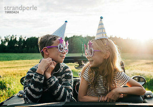 Mädchen und Junge saßen mit Partyhüten und Brillen in einem Cabrio