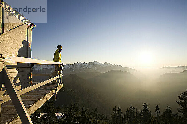 Eine junge Frau steht auf der Veranda eines Feuerwachturms mit Blick auf weite Bergketten.