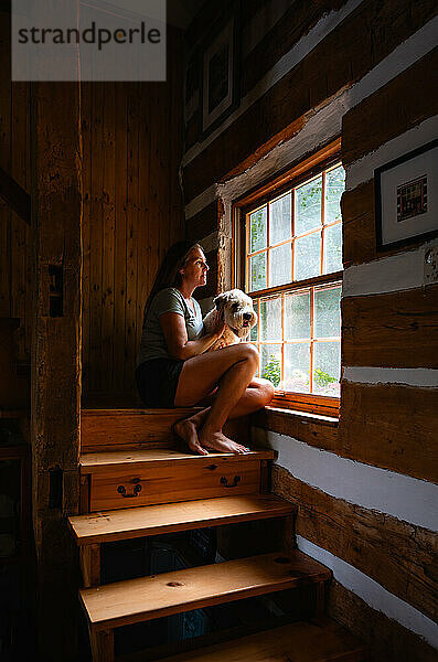 Frau und Hund sitzen neben einem Fenster auf der Treppe eines Blockhaushauses.