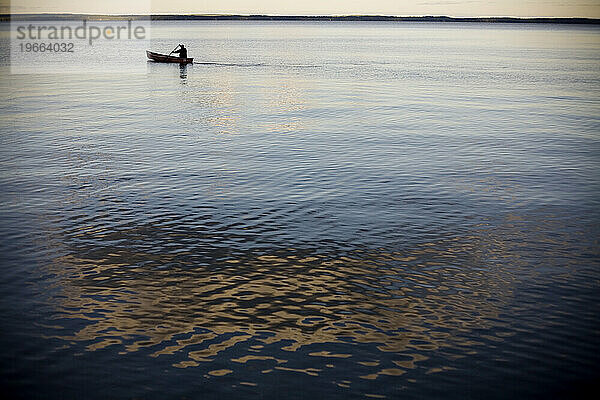 Mit der Spiegelung einer Wolke im ruhigen  blauen Wasser treibt ein Mann friedlich seine Silhouette auf dem glatten Wasser ab