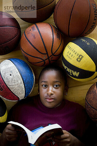 A young boy lies amongst a group of basketballs while reading a book.