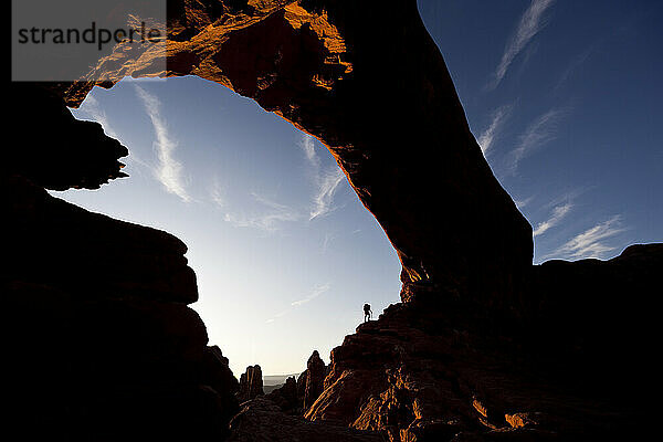 Silhouette eines Wanderers  der unter einem natürlichen Felsbogen im Arches-Nationalpark in Utah steht.