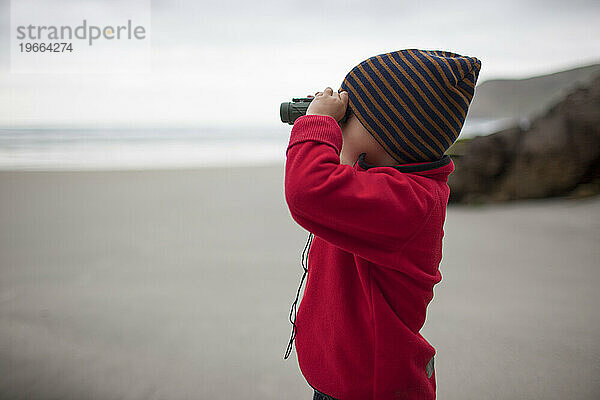 Kleiner Junge schaut durch ein Fernglas an einem Strand in Neuseeland
