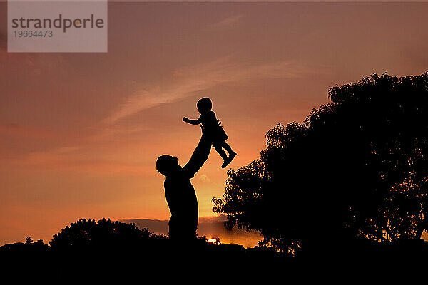 Die Silhouette des Vaters hebt seinen Sohn hoch vor einem Sonnenuntergangshimmel.