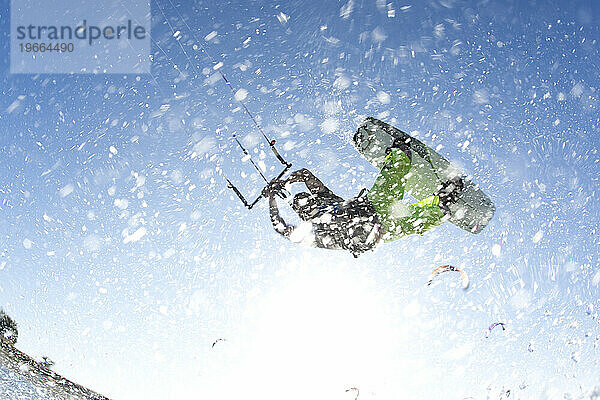 Tiefwinkelperspektive eines Kiteboarders  der mit viel Gischt in der Luft in die Sonne springt.