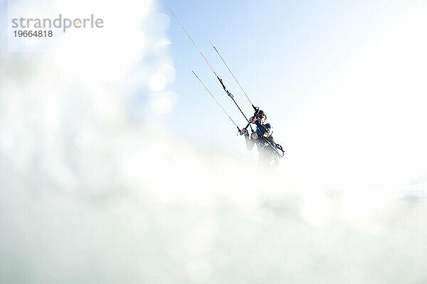 Tiefwinkelperspektive eines Kiteboarders  umrahmt von ausgeblasenem Wasser und Himmel.