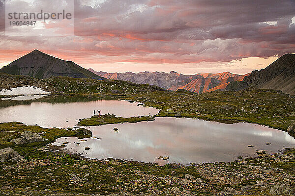Zwei Menschen stehen bei Sonnenuntergang am Rande eines Sees in den Bergen.