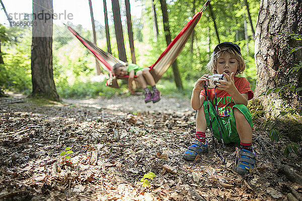 Ein 5-jähriges Mädchen in einer Hängematte  während ihr Bruder mit einer Kamera spielt. In einem Wald  Frankreich.