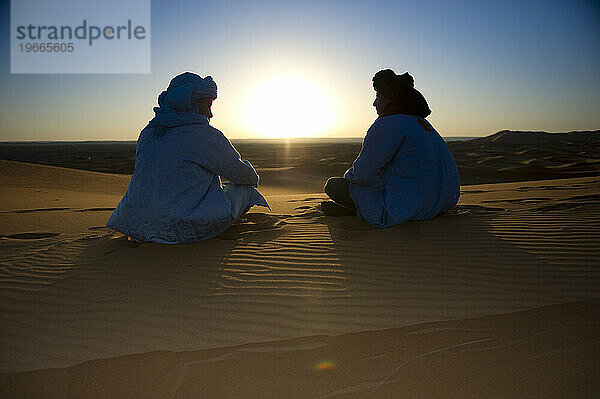 Zwei Jungen in traditioneller nordafrikanischer Kleidung sitzen bei Sonnenaufgang auf einer Sanddüne.