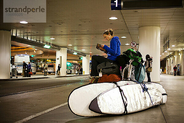 Eine junge Frau wartet auf eine Flughafenfahrt  während sie nachts auf ihren Taschen neben ihren Surfbrettern am Flughafen sitzt.