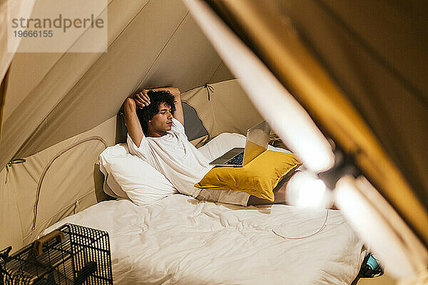 Ein lateinamerikanischer Mann schaut sich eine Serie auf dem Computer an und liegt auf dem Bett in einem Zelt