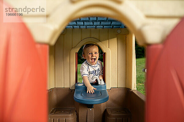 Kleinkind steckt lachend seinen Kopf durch das Fenster des Spielhauses
