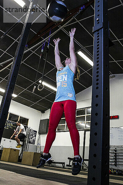 Ein Mädchen macht während eines Crossfit-Workouts einen Wallball  während im Hintergrund ein Mann Boxjumps macht.