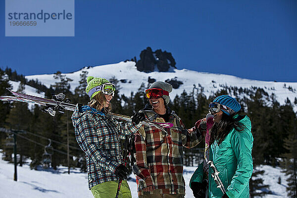 Drei Freunde (ein Mann und zwei Frauen) hängen mit Skiern und einem Snowboard vor einem Skigebiet herum.