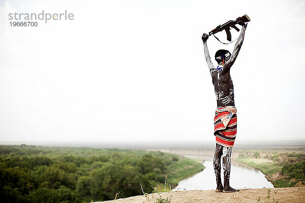 Ein junger Mann hält sein Kalaschnikow-Gewehr über seinen Kopf und blickt dabei auf den Omo-Fluss im abgelegenen Omo-Tal in Äthiopien.