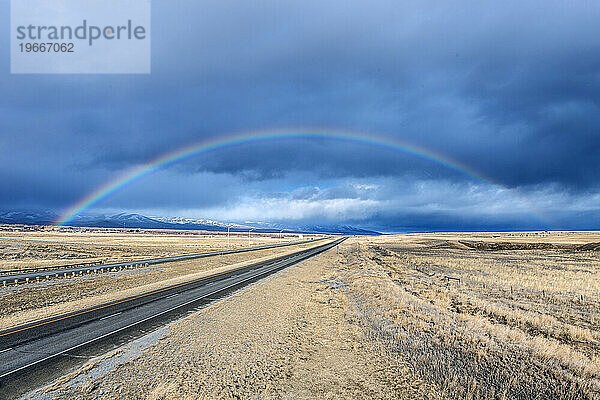 Eine bis zum Horizont reichende Autobahn durch eine flache Landschaft  ein Regenbogen über der Straße.