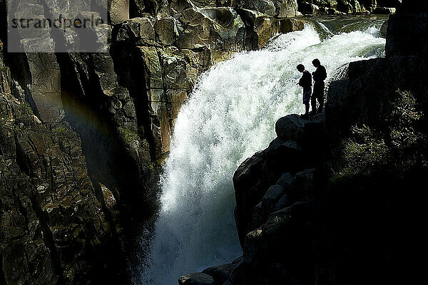 Silhouette zweier Kajakfahrer  die oben auf einem großen Wasserfall mit einem Nebelregenbogen stehen.