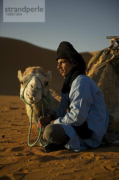 A camel guide sits with his animal in the sand dunes at sunrise.