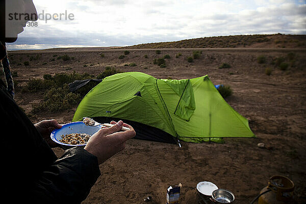 Ein Mann isst Haferflocken  nachdem er vom Camping in der Wüste aufgewacht ist.